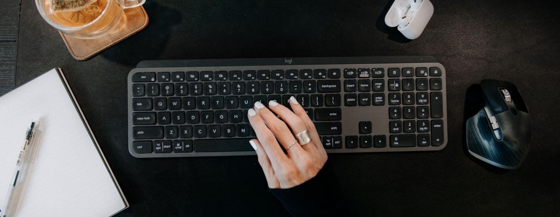 woman's hand on a keyboard surrounded by office items and a cup of tea on a coaster